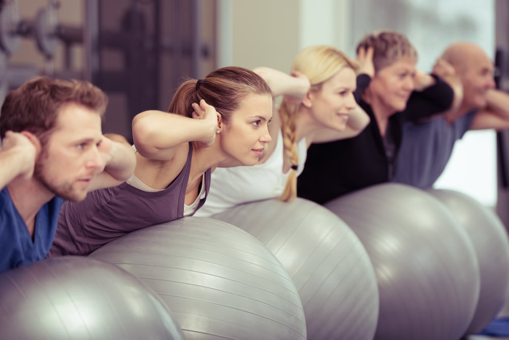 Group of diverse people in a receding line doing pilates in a gym balancing over the gym balls with their hands laocked behind their necks toning their muscles